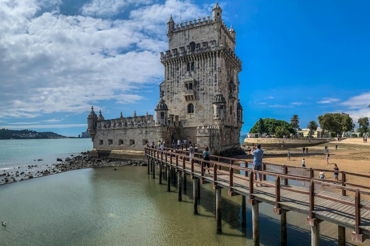 Historic stone tower by water with footbridge, people walking, and a partly cloudy sky.