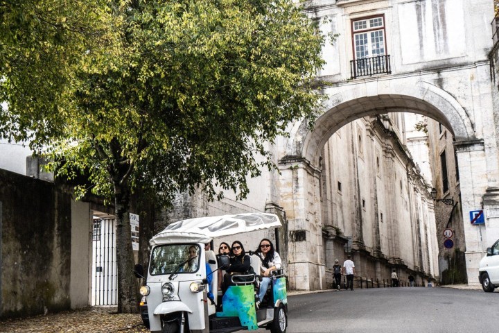 A tuk-tuk with three passengers drives down a street near an archway and large building.