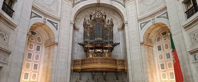 Ornate organ on balcony in a grand neoclassical church interior with Portuguese flag.