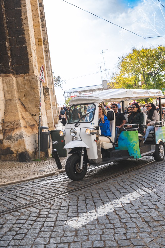 White tuk-tuk with passengers driving on a cobblestone street near a large stone structure.