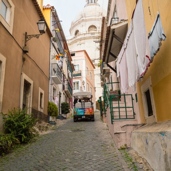 Narrow cobblestone street between buildings with a small vehicle and laundry hanging, dome visible in background.