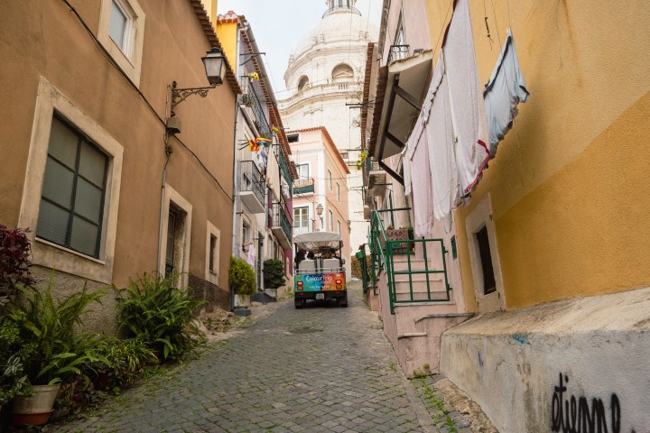 Narrow cobblestone street between buildings with a small vehicle and laundry hanging, dome visible in background.