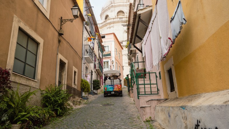 Narrow cobblestone street between buildings with a small vehicle and laundry hanging, dome visible in background.