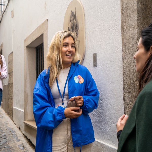 Woman in blue jacket talking to another person on a narrow cobblestone street.