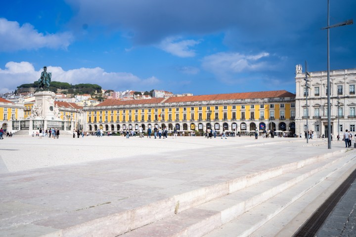 Sunny square with a statue, yellow building, and people walking, against a blue sky.