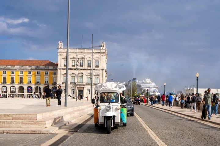 Urban scene with a tuk-tuk driving on a cobblestone street, people walking, and historic buildings nearby.