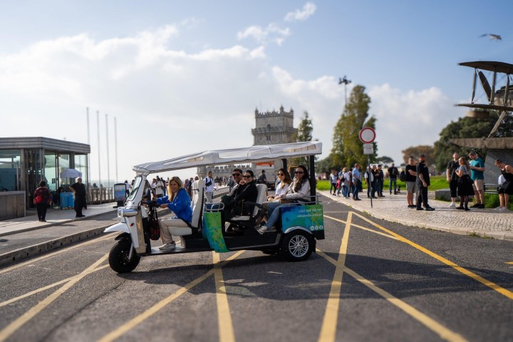 Tourists on a tuk-tuk near a historic tower with a crowd in the background.