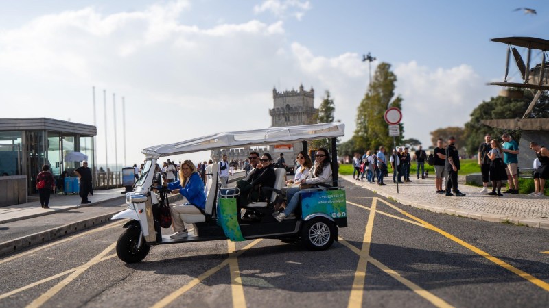 Tourists on a tuk-tuk near a historic tower with a crowd in the background.