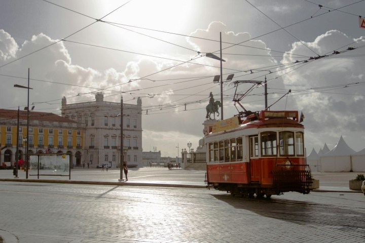 Vintage red tram in a sunlit European square with historic buildings and a statue.