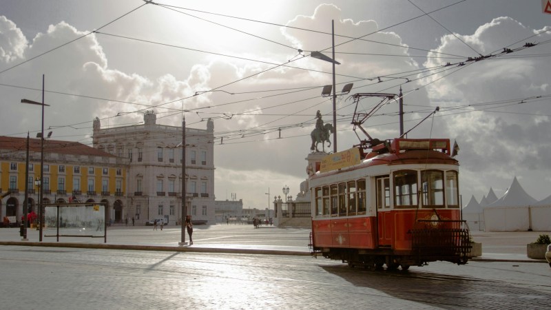 Vintage red tram in a sunlit European square with historic buildings and a statue.