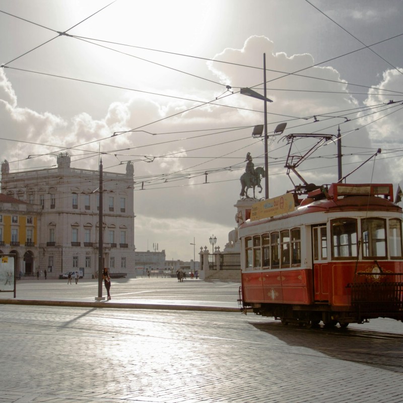 Vintage red tram in a sunlit European square with historic buildings and a statue.