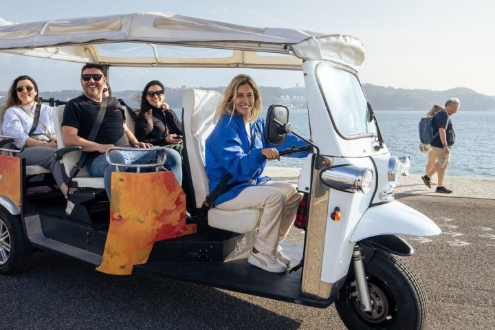 Four people smiling on a tuk-tuk by the waterfront on a sunny day.