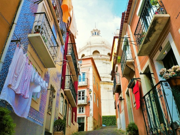 Narrow colorful street with laundry hanging, leading to a dome in the background.